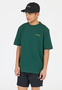 Vihreä puuvillapaita, jossa on lyhyet hihat ja pyöreä kaula-aukko. Paidassa on pieni kultainen "LES' ARCS" -logo rinnassa. Musta lippalakki, jossa on brodeeraus.