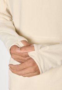 Cream-colored fleece top with a relaxed fit and thumbholes in the sleeves, designed for comfort and warmth, featuring soft texture.