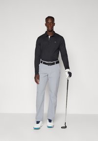 Μαύρο πόλο μπλουζάκι μακριά μανίκια, ανοιχτό γκρί παντελόνι, μαύρη ζώνη, γάντι γκολφ, άσπρα παπούτσια με μπλε λεπτομέρειες. Στέκεται με ένα κλάμπ γκολφ.