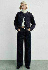 Temno denim kratka jakna z gumbi in sprednjimi žepe, v kombinaciji z visokim pasom temnimi kavbojkami. Pod njo je bela turtlenecka majica.