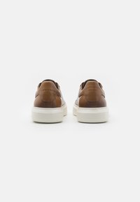 Zapatillas de cuero marrón con un acabado liso, suela de goma blanca y detalles de costura sutiles. Vista trasera que muestra el collar acolchado y el logo.