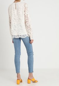 Top in pizzo bianco con maniche lunghe, dotato di apertura a chiave sul retro, abbinato a jeans azzurri strappati e sandali con tacco a blocco gialli.
