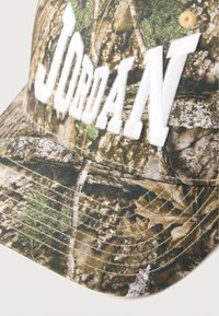 Căciulă camuflaj cu broderie albă "JORDAN". Prezintă nuanțe de verde și maro, material texturat și detalii vizibile de cusătură.
