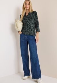 Haut vert foncé avec imprimé léopard, manches trois-quarts, associé à un jean large bleu et des baskets blanches. Sac beige sous le bras.
