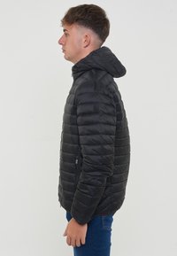 Giubbotto puffer nero con trapuntatura orizzontale, chiusura con zip, cappuccio regolabile e tasche con zip, indossato con jeans blu.