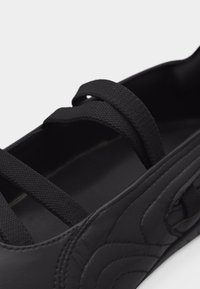 Chaussure noire de style Mary Jane avec deux sangles élastiques croisées et un détail matelassé sur le côté.