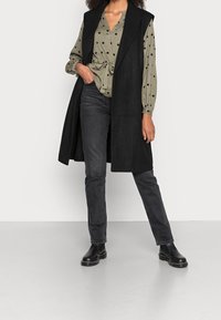 Un long gilet noir sur un chemisier vert à pois avec ceinture à la taille, associé à un jean noir et des bottines noires. Texture lisse et design simple.