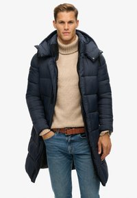 Marinepufferjakke med hætte, der har quiltet tekstur, en fuld lynlås og sidelommer. Båret over en beige sweater og jeans, accessoriseret med et bælte.