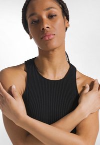 Sort ribbet tanktop med høj hals. Modellen krydser armene og fremviser stoftekstur og tætsiddende design uden yderligere detaljer.