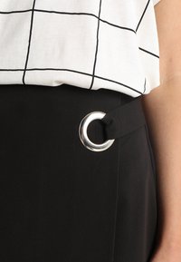 Pantaloni neri a vita alta con dettaglio in argento a forma di cerchio sulla cintura, abbinati a una maglietta bianca con motivi a quadri neri.