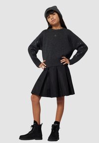 Czarny sweter z dzianiny z cekinowymi detalami, czarna plisowana spódnica z geometrycznym wzorem oraz czarne buty na sznurówki na modelce-dziecku.