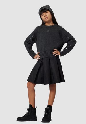 Zwarte gebreide trui met pailletten, zwarte geplooide rok met geometrisch patroon, en zwarte veterschoenen op een kindermodel.