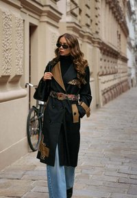 Czarny dwurzędowy płaszcz trencz z brązowymi akcentami i kieszenią w zwierzęcy wzór. Połączony z jasnoniebieskimi dżinsami i dodatkami.