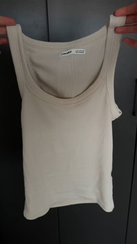 Cremfarbener, gerippter Tanktop mit breiten Trägern, rundem Ausschnitt und leicht gewölbtem Saum. Das Etikett trägt die Aufschrift "Pull&Bear", hergestellt in Bangladesch.