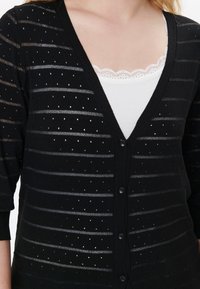 Cardigan nero con design a strisce orizzontali e piccoli punti. Presenta un colletto a V, chiusura con bottoni ed è sovrapposto a una maglia bianca con orli in pizzo.