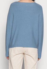 Osoba w luźnym, jasnoniebieskim swetrze dzianinowym i off-white spodniach z korduroju, widoczna od tyłu na neutralnym tle.