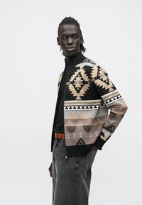 Črno-bež vzorčast kardigan z geometrijskimi vzorci, visok ovratnik, zadrga, nošen čez črno srajco in sive kavbojke.