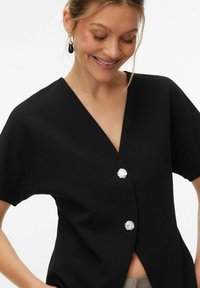 Must lühikeste varrukatega top, millel on V-kaelus ja kaks dekoratiivset hõbedast nuppu. Ühtlane tekstuur ja lõdvestunud lõige. Minimalistlik disain, ilma mustriteta.