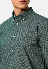 Camisa verde de botones con un patrón a cuadros, presenta un cuello abotonado y un pequeño parche con logotipo en el pecho. Tejido de algodón suave.