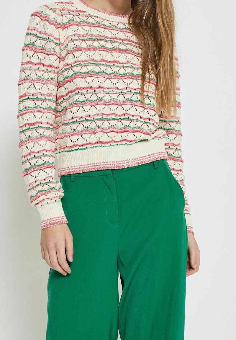 Femme portant un pull en maille à motifs rose, vert et blanc avec un pantalon vert taille haute, debout, les mains détendues le long du corps.