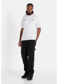 Camiseta blanca de algodón con un logo en la parte frontal, combinada con pantalones cargo negros que cuentan con bolsillos y acentos metálicos. El modelo está de pie sobre un fondo neutro.