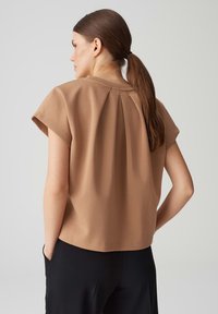 Top marrone a maniche corte con scollatura rotonda e dettaglio a pieghe sul retro, abbinato a pantaloni neri. Tessuto morbido e liscio.