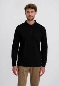 Mann mit kurzen lockigen Haaren und Bart, der ein schwarzes Polohemd mit langen Ärmeln und beigefarbene Cordhosen trägt und vor einem neutralen Hintergrund steht.