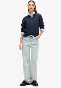 Sudadera azul marino con cuello de cremallera sobre una camisa azul clara, combinada con jeans azules claros y zapatos negros, destacando una hebilla de cinturón plateada.