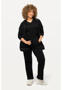 Černá mikina na zip s tkaničkami, černé tričko a černé kalhoty. Model nosí bílé tenisky, má kudrnaté vlasy. Hladká textury po celém povrchu.