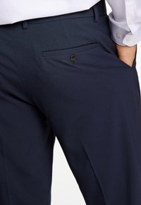 Pantaloni blu navy in tessuto strutturato, dotati di una tasca posteriore singola con chiusura a bottone e design frontale piatto.