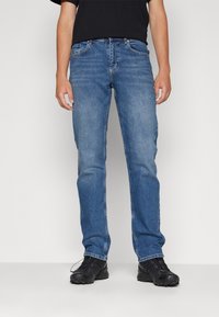 Vaqueros de mezclilla azul clásico con un corte recto, que cuentan con cinco bolsillos, cremallera y pasadores para cinturón. Se llevan con calzado negro.