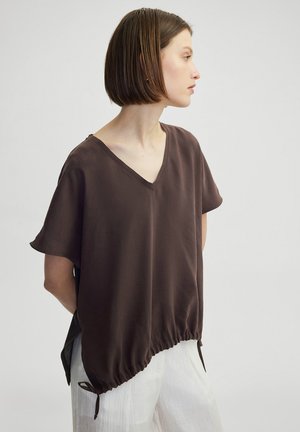 Brown, loose-fitting V-neck top with short sleeves and gathered hem detail. Smooth texture with side slits for ease of movement.