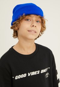 Zila adīta beanie un melna džemperis ar tekstu "TIKAI LABAS ENERĢIJAS". Džemperim ir apaļa kakla izgriezums un garas piedurknes ar baltām akcentiem.