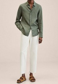 Grünes Leinenhemd mit Knöpfen, weiße locker sitzende Hose und braune Sandalen. Das Outfit zeichnet sich durch eine entspannte Passform und ausgefranste Hosensäume aus.