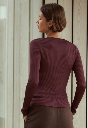 Femme aux cheveux bruns courts portant un haut bordeaux côtelé à manches longues ajusté et un pantalon marron, debout, les mains dans les poches, tournée vers la gauche.