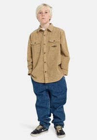 Camicia in corduroy color nocciola con due tasche sul petto e dettaglio con logo, abbinata a jeans in denim blu larghi e sneakers nere.