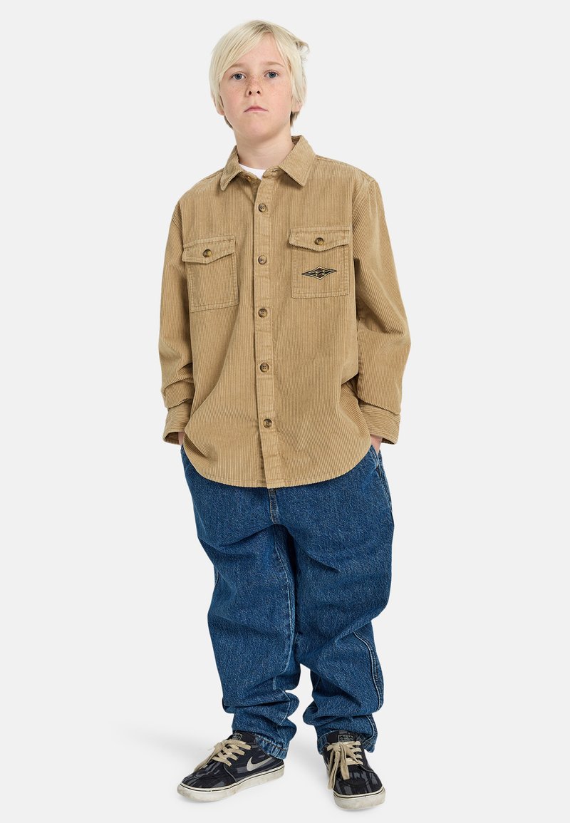 Camicia in corduroy color nocciola con due tasche sul petto e dettaglio con logo, abbinata a jeans in denim blu larghi e sneakers nere.