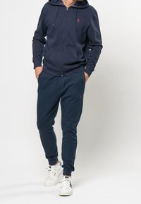 Marinblå zip-up hoodie med kenguruficka och röd logga, kombinerad med matchande joggerbyxor med resår i midjan och muddar.