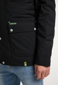 Veste noire avec une poche dotée de boutons-pression argentés et une étiquette logo verte. Tissu texturé avec une finition lisse sur les manches.