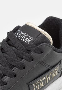 Svart lädersneaker med strukturerad yta, snörningsdesign, logotyp med guldfinish och vit gummisul. Har en vadderad krage.