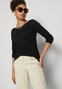 Haut noir à manches longues avec un décolleté bateau, associé à un pantalon large crème. Les accessoires incluent des lunettes de soleil marron surdimensionnées et un collier en chaîne.