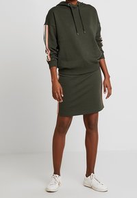 Un hoodie vert foncé avec des rayures roses et blanches sur les manches, associé à une jupe verte foncée assortie. Des baskets blanches complètent la tenue.