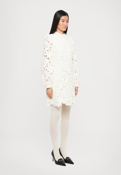 Witte kantjurk met bloemenuitsparingen, hoge kraag en pofmouwen, gecombineerd met lichtbeige panty’s en zwarte puntige hakken met een gouden accent.