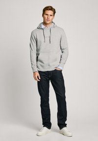 Sudadera gris con un bolsillo frontal y capucha con cordón, superpuesta a una camisa de color azul claro con cuello, combinada con jeans oscuros y zapatillas blancas.