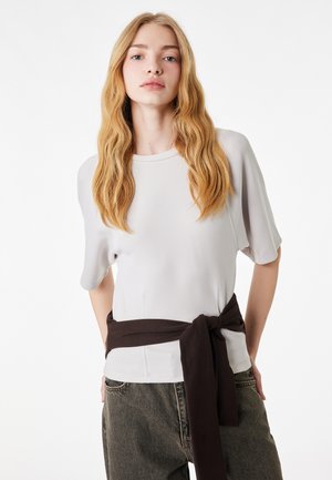 Junge Frau mit langen, welligen blonden Haaren, die ein hellgraues Kurzarmoberteil und dunkle Hosen trägt, mit einem schwarzen Pullover um die Taille gebunden.