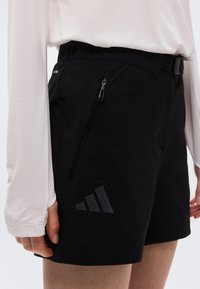 Άτομο που φοράει μαύρο σορτς με κεκλιμένη τσέπη με φερμουάρ και διακριτικό λογότυπο, συνδυασμένο με λευκή μπλούζα μακριάς μανίκας, εμφανιζόμενο από τη μέση έως το μέσο του μηρού.