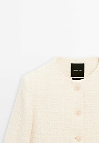 Veste texturée crème avec un col rond et trois boutons, avec une étiquette noire indiquant Massimo Dutti.