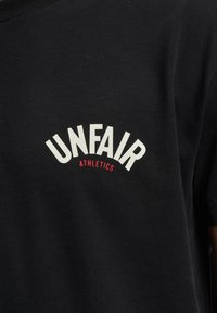 Schwarzer Stoff mit weißem Text „UNFAIR“, der über kleinerem rotem Text „ATHLETICS“ auf einem Kleidungsstück, wahrscheinlich einem T-Shirt, gebogen ist.
