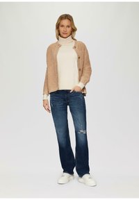 Jasno beżowy sweter z dzianiny z guziczkami, noszony na kremowym golfie. Połączony z niebieskimi dżinsami o prostym kroju z przetarciami i białymi trampkami.