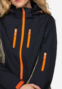 Giacca impermeabile blu navy e verde oliva con dettagli arancioni sulla cerniera, quattro tasche con zip, colletto alto e polsini regolabili.